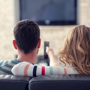 Two people sit on a couch watching TV; one holds a remote, arm wrapped around the other in a cozy living room setting.