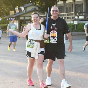 Participants smile while holding a lightsaber, walking in a race event near a traditional Asian building. Shirts read 