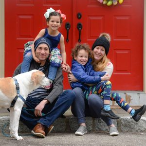 Family sitting joyfully; children on parents' laps, dog standing nearby; posed in front of vibrant red double doors.