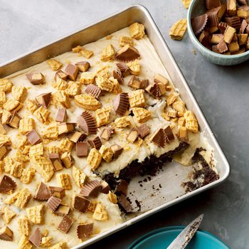 Baking tray displays chocolate cake topped with peanut butter cups and cookies; a slice is missing. Bowl with toppings nearby on gray surface.