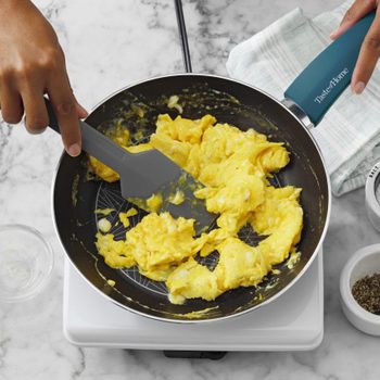 Scrambled eggs cooking in a pan on a stove, surrounded by hands holding utensils, salt and pepper shakers, and a towel on marble.