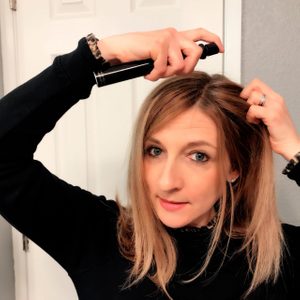 A woman applies hair product, holding a spray bottle over her head, in a bathroom with a white door in the background.