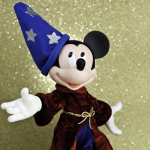 Character wearing wizard hat, extending arm, stands against gold sparkly background.