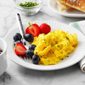 Scrambled eggs with chives sit on a plate with blueberries and strawberries, surrounded by a coffee cup, toast, and a napkin on a marble table.