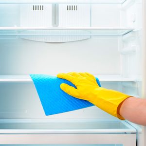 A gloved hand cleans an empty refrigerator shelf with a blue cloth in a bright kitchen setting.