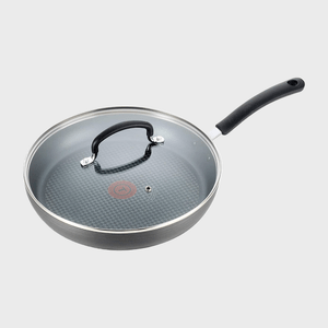 A frying pan with a glass lid rests on a plain background, featuring a black handle and a red circle at the center of the surface.