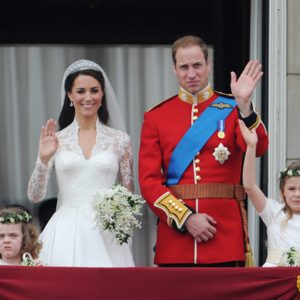 Bride and groom wave from balcony, accompanied by three children with flower crowns, in a celebratory outdoor setting.