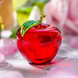 A red glass apple sits on a reflective surface, surrounded by blurred pink and green shapes, suggesting flowers and leaves.