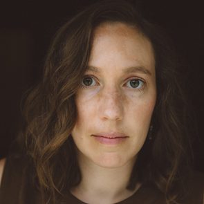 A woman gazes directly, her face framed by wavy hair, set against a dark background.
