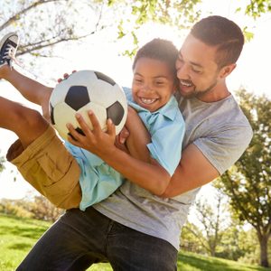 Child holds soccer ball, smiling, while being lifted by a man in a sunny park, surrounded by trees.