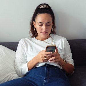 Person using a smartphone, lounging on a gray couch with white pillows, wearing a white shirt and blue jeans, indoors.