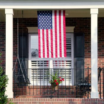American flag hangs from a porch with brick walls, flanked by a swing chair and potted flowers, under a roof supported by columns.