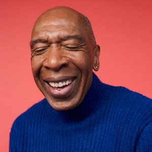 Smiling person wearing a blue sweater against a solid red background, eyes closed, expressing joy.