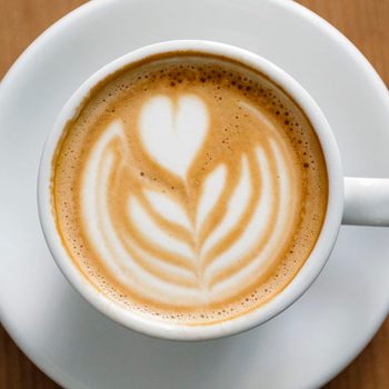A white coffee cup displays heart-shaped latte art, sitting on a matching saucer on a wooden table.