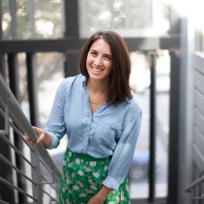 A smiling person ascends stairs, holding the railing, in a well-lit, modern building with metal and glass elements.