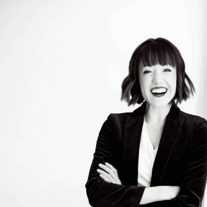 A person smiles widely, crossing their arms, in a brightly lit space, wearing a blazer and white shirt.