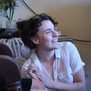Person smiling, sitting indoors beside a table with flowers, a phone, and a crocheted cloth, looking towards the right.