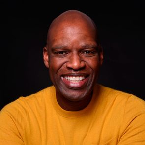 Man smiling warmly against a dark background, wearing a yellow sweater.