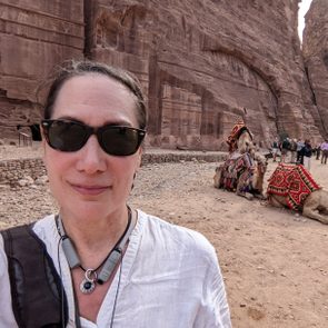 Person wearing sunglasses smiles outdoors; camels rest in the background with colorful blankets near tall rock formations, several people standing nearby.
