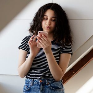 A person wearing a striped shirt uses a smartphone, standing against a white wall with diagonal light shadows.