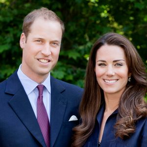 Two people stand smiling, dressed formally with a suit and blouse, against a lush green leafy background outdoors.