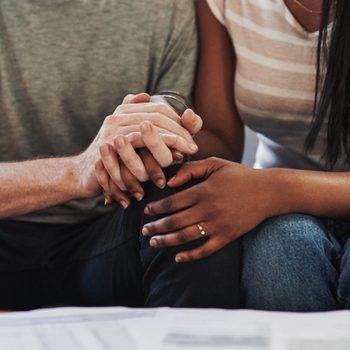 Hands clasp together supportively; two people sit closely on a sofa, with documents visible on a table in front.