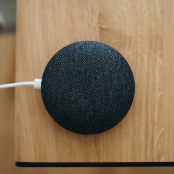 A small, round smart speaker rests on a wooden surface, plugged in with a white cable, near the edge of a tabletop.