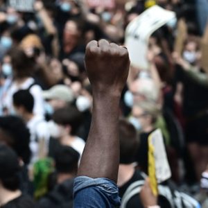 A raised fist stands out amidst a dense crowd of masked protesters, many holding signs, during a demonstration.