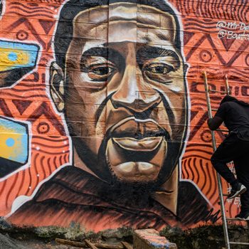 Mural depicts a man's face with intricate patterns. Artist climbs ladder nearby, surrounded by text highlighting themes of police brutality and killings.