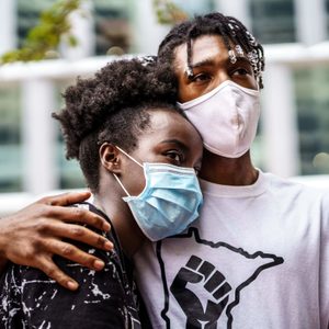 Two people wearing masks embrace closely, standing outside in front of a modern building with large windows.