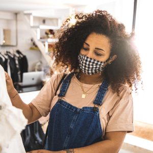 Person wearing a checkered mask examines clothes on a rack in a bright, modern store environment.