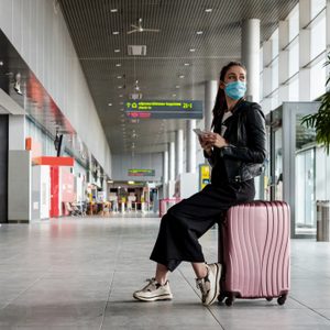 Woman wearing a mask, sitting on pink suitcase, holds phone in a spacious airport terminal with signs and seating areas in the background.
