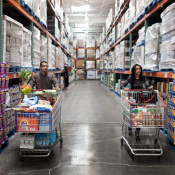Two shoppers push full carts through a large warehouse store aisle lined with shelves of bulk products.