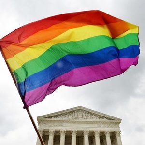 Rainbow flag waves in front of a neoclassical building with tall columns under a cloudy sky.