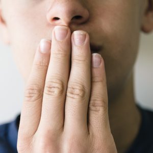 Hand covering mouth, fingers aligned vertically, against a blurred face with neutral expression in soft lighting.