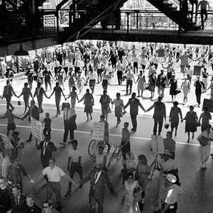 Protesters hold hands, forming a large circle, under a bridge, carrying signs.