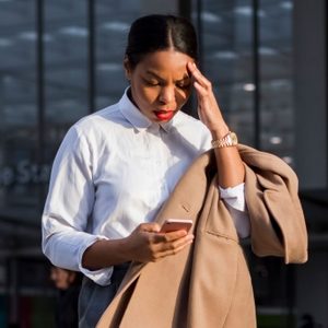 A person looks at a phone, holding a coat, appearing concerned, in a modern, urban outdoor setting.