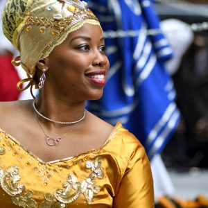 Woman smiling, wearing ornate yellow outfit and jeweled headscarf, surrounded by people and colorful fabrics in festive setting.