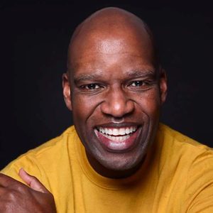 Man smiling broadly, wearing a yellow shirt, against a dark background.