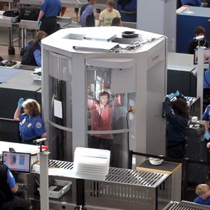 Person inside a body scanner standing with arms raised; airport security personnel monitor screens and assist nearby passengers in a busy terminal setting.