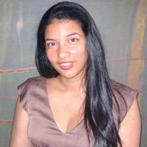 A person with long, dark hair smiles softly, standing against a subtly patterned background.