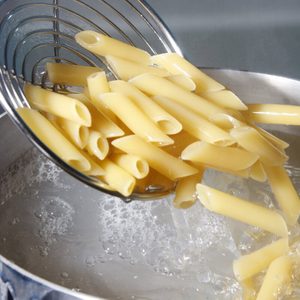 Pasta drains from a metal strainer above a pot of boiling water in a kitchen setting.