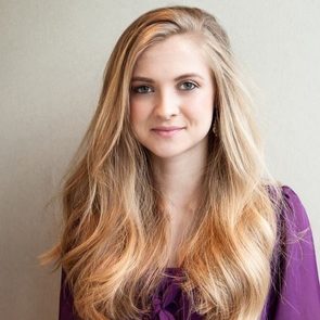A person with long, wavy blonde hair smiles slightly, wearing a purple top, posing against a plain light background.