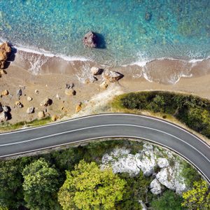 A winding road curves beside a sandy beach with rocks, adjacent to turquoise water and bordered by lush greenery.