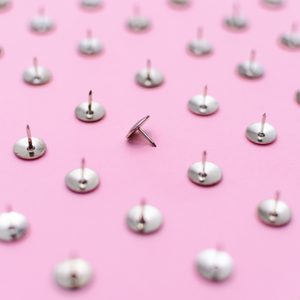 A single thumbtack leans among many upright thumbtacks on a pink surface, creating a patterned effect.
