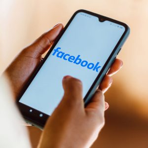 Hands hold a smartphone displaying the Facebook logo on the screen, in a softly lit indoor environment.