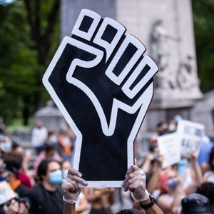 Raised fist sign held up at a protest in a park, surrounded by people wearing masks and holding signs.