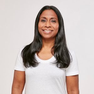 A person smiles while standing, wearing a white shirt, against a plain, light-colored background.