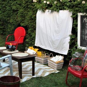 Screen hangs with movie showtimes written nearby; chairs, a blanket, a table with a small fire pit, and popcorn baskets surround it.