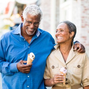 Two people smile, walking with ice cream cones, in an outdoor urban area with blurred buildings in the background.